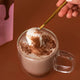 A clear glass mug filled with hot chocolate topped with white foam and cocoa powder, with a hand holding a gold spoon dipping into the foam.