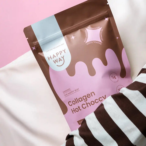 A stand-up pouch of Happy Way Collagen Hot Choccy lies on a pink and white surface with a brown-and-white striped fabric partially visible at the bottom right.