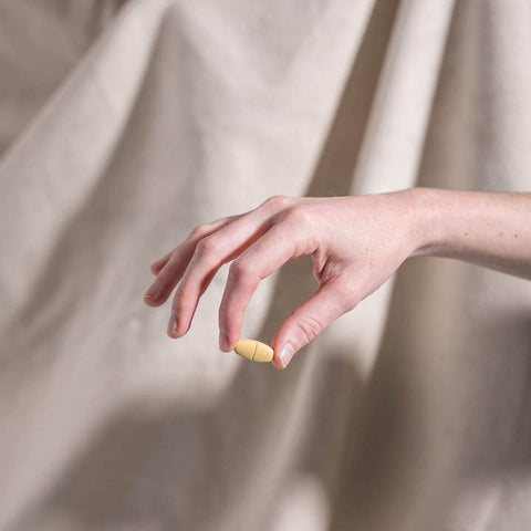 Hand holding a yellow pill against a beige curtain background