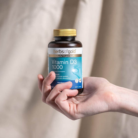 Hand holding a bottle of Herbs of Gold Vitamin D3 1000 against a neutral background.