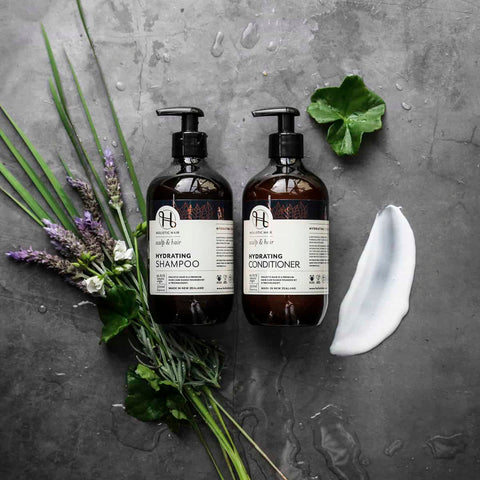 Two bottles of shampoo and conditioner with herbs and a leaf on a gray surface.