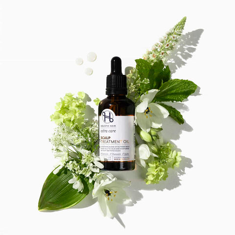 Bottle of hair treatment oil surrounded by green leaves and white flowers on a white background.