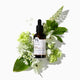 Bottle of hair treatment oil surrounded by green leaves and white flowers on a white background.