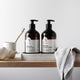 Two bottles of sensitive shampoo and conditioner on a wooden surface with a towel and bowl in the background.