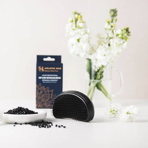 Black hairbrush with a bowl of black hair products and a vase of white flowers on a light background.