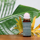 Honest Skincare Organic Castor Oil Rose Quartz Roller 50ml lifestyle shot of the bottle with its rose quartz roller beside large green tropical leaves