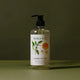 A bottle of hand wash with a black pump dispenser and a white label featuring botanical illustrations, set against a dark olive green background.