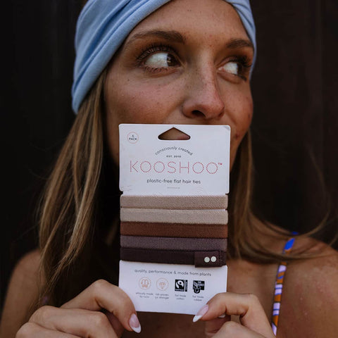 Woman holding a package of Kooshoo Plastic-Free hair ties against her face with a dark background.