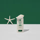 A baby sunscreen stick on a white surface against a dark green background, styled with a white seashell on top of the stick and a white starfish prop on the left.