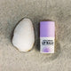 A small tube of lip balm with a purple cap on sand next to a white clam shell.