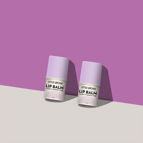 Two small lip balm tubes standing upright on a light gray surface against a purple background. The tubes are pale lavender with white labels.