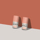 Two lip balm tubes standing upright on a light gray surface against a terracotta background. The tubes are pale peach-pink with white labels.