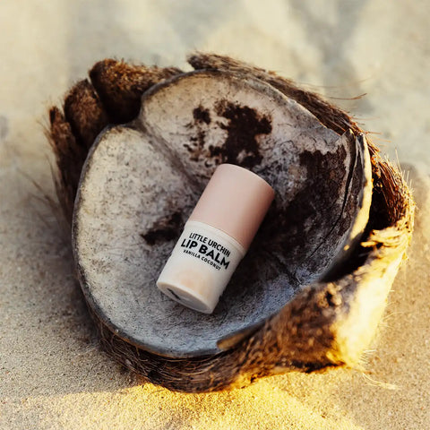A small lip balm stick labeled 'Little Urchin Lip Balm' rests on a cut coconut shell placed on sandy ground outdoors.
