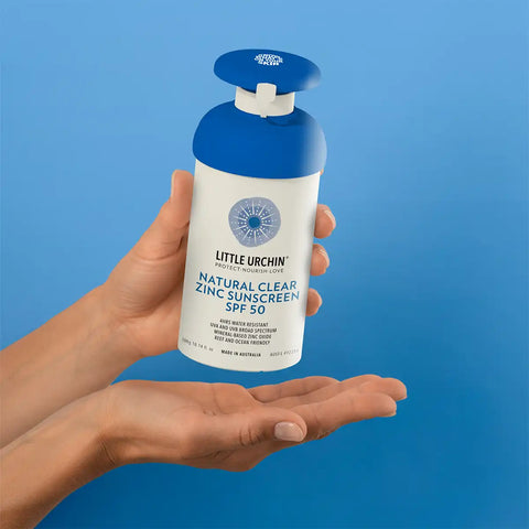 A person's hands holding a bottle of Little Urchin Natural Clear Zinc Sunscreen SPF 50 against a solid blue background.