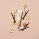 A tube of Little Urchin Vanilla lip balm is arranged on a soft pink background with vanilla beans, small white flowers, and the cap placed beside it.