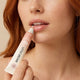 A close-up of a woman with red hair applying a peachy-pink lip balm from a Little Urchin tube to her lips.