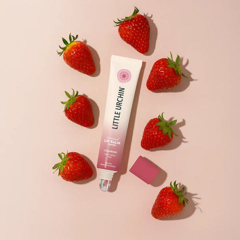 A tube of Little Urchin Strawberry Lip Balm is arranged on a light pink background, surrounded by fresh strawberries.
