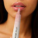 Close-up of a person applying a pink lip balm from a white and pink tube. The product is shown being applied to the lips against a clean skin background.