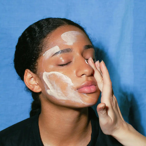 A close-up of a person with white cream or makeup applied to their face, applying it with their hand against a solid blue background.