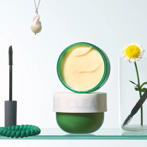 An open round cosmetic jar with a pale yellow cream product inside and a green rim, styled on a light surface with a mascara wand, a green cord, a pendant necklace, and a daisy in a glass vase.
