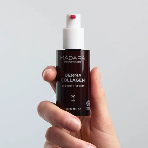 A hand holding a dark brown bottle of Mádara Organic Ingredients Derma Collagen Peptides Serum against a light grey background.