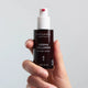 A hand holding a dark brown bottle of Mádara Organic Ingredients Derma Collagen Peptides Serum against a light grey background.