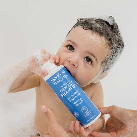 Baby holding a bottle of MooGoo Natural Gentle Shampoo with soap suds on a white background