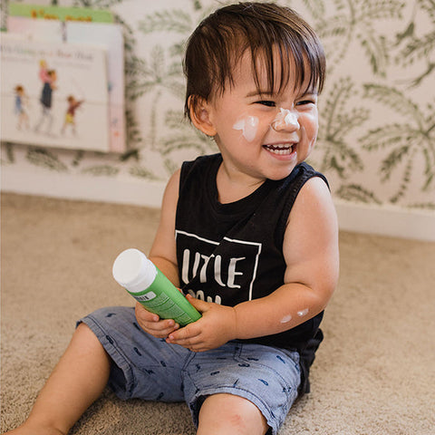 Child holding a green bottle with a white cap, sitting on a carpeted floor, with moisturiser all over his face.