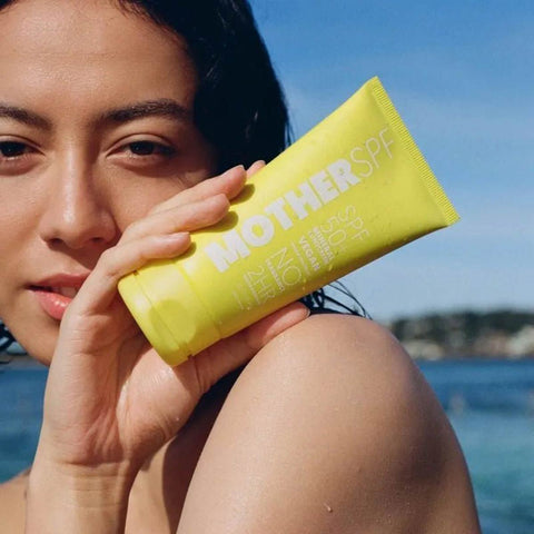Person holding a yellow 'MOTHERSPF' sunscreen tube by the water.