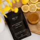 MUD Organics detox supplement powder package on a wooden surface with lemons and a glass of detox powder in water/juice.