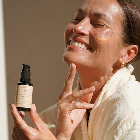 A woman in a white robe smiles while holding a small black skincare bottle labelled MUKTI.