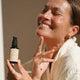 A woman in a white robe smiles while holding a small black skincare bottle labelled MUKTI.