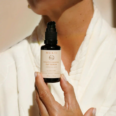A person in a white robe holds a small black pump bottle labeled MUKTI Bioactive Renewal Day Serum against a softly lit neutral background.