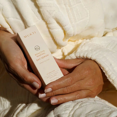 A person holding a cream-colored box of MUKTI Bioactive Renewal Day Serum, wrapped in a textured white robe.