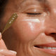 Close-up of a woman applying cream from a gold spatula labelled MUKTI onto her cheek.