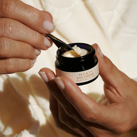 A close-up of hands holding a small black jar of MUKTI Eye Crème and scooping cream with a small spoon.