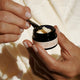 A close-up of hands holding a small black jar of MUKTI Eye Crème and scooping cream with a small spoon.