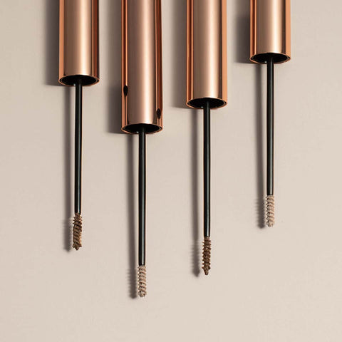 Four rose gold mascara tubes with black threads on a beige background.