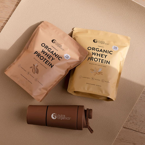 Two packages of Nutra Organics Organic Whey Protein range & a shaker bottle on a wooden surface.
