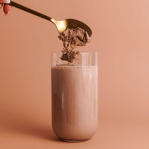Spoon adding Nutra Organics Organic Whey Protein - Chocolate 750g powder to a glass of milk on a peach background.