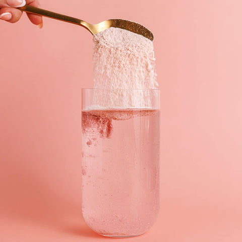 Gold spoon pouring a white powdery substance into a glass of pink liquid on a pink background.