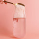 Gold spoon pouring a white powdery substance into a glass of pink liquid on a pink background.