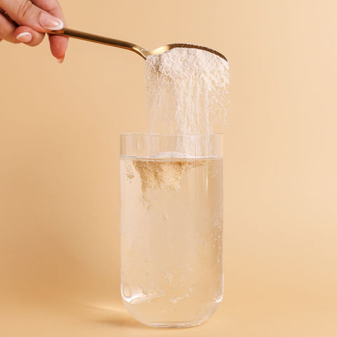 Spoon pouring a white powder into a glass of water on a beige background.