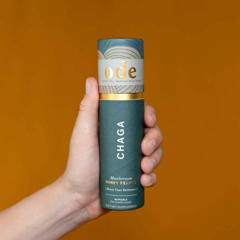 Hand holding tube of Chaga Mushroom Honey Pearls against an orange background. 