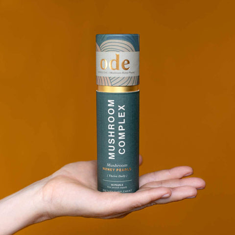 Hand holding tube of Mushroom Complex Honey Pearls against an orange background. 