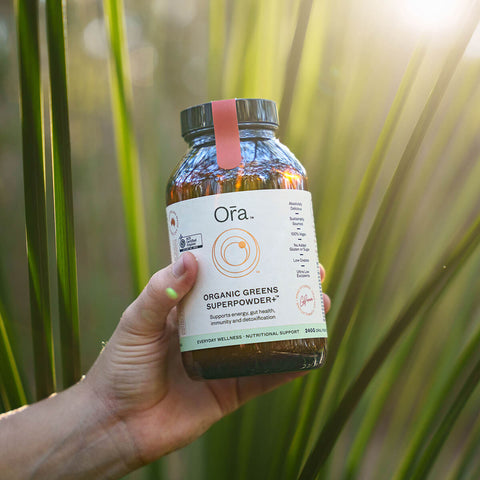 Hand holding 240g jar Ora Organic Greens Superpowder+, vegan supplement to support energy, gut health, immunity, and detox.