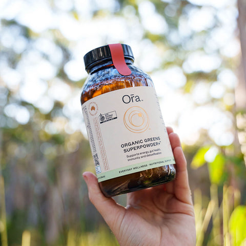 Hand holding 240g jar Ora Organic Greens Superpowder+, vegan supplement to support energy, gut health, immunity, and detox.