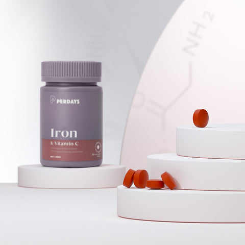 Iron & vitamin C tablet bottle styled on a white cylindrical surface with a stack of design elements with multiple reed tablets on top.
