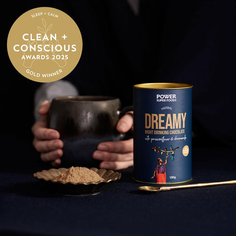 Can of Power Super Foods Dreamy Night Drinking Chocolate with a spoonful of chocolate powder & a cup on a dark background.