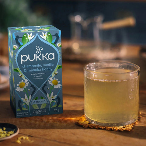 Pukka chamomile, vanilla, and manuka honey tea box with a glass of tea on a wooden surface.